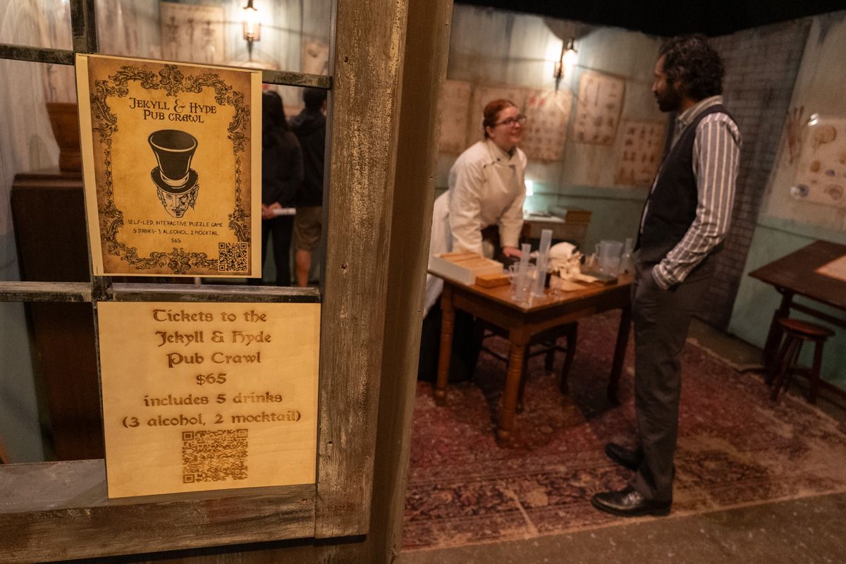 A man wearing a vest and a pinstriped shirt  confers with a woman in a white lab coat in a room decorated like a Victorian laboratory, with signs on the doorway in the foreground displaying information about the "Jekyll and Hyde Pub Crawl."