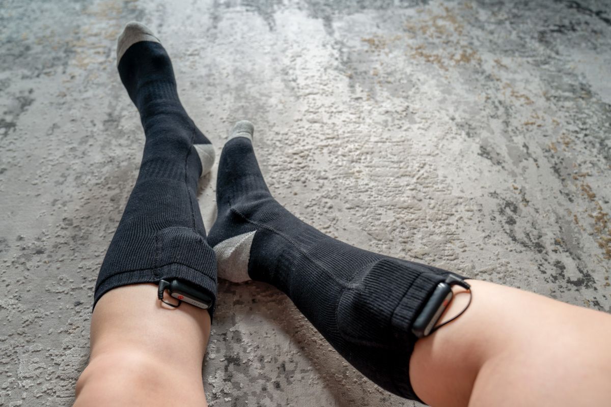 Point of view of a pair of legs and feet wearing black heated ski socks with battery packs, sitting against a textured grey surface.