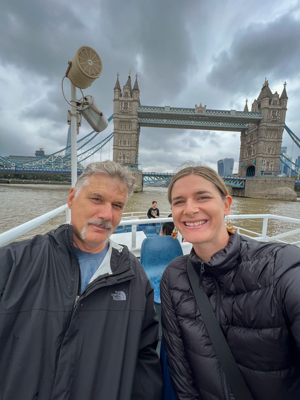An older man in a black jacket poses next to a young woman in a black Lightweight Down Puffer Jacket from the deck of a boat in front of the London Bridge on a overcast day.