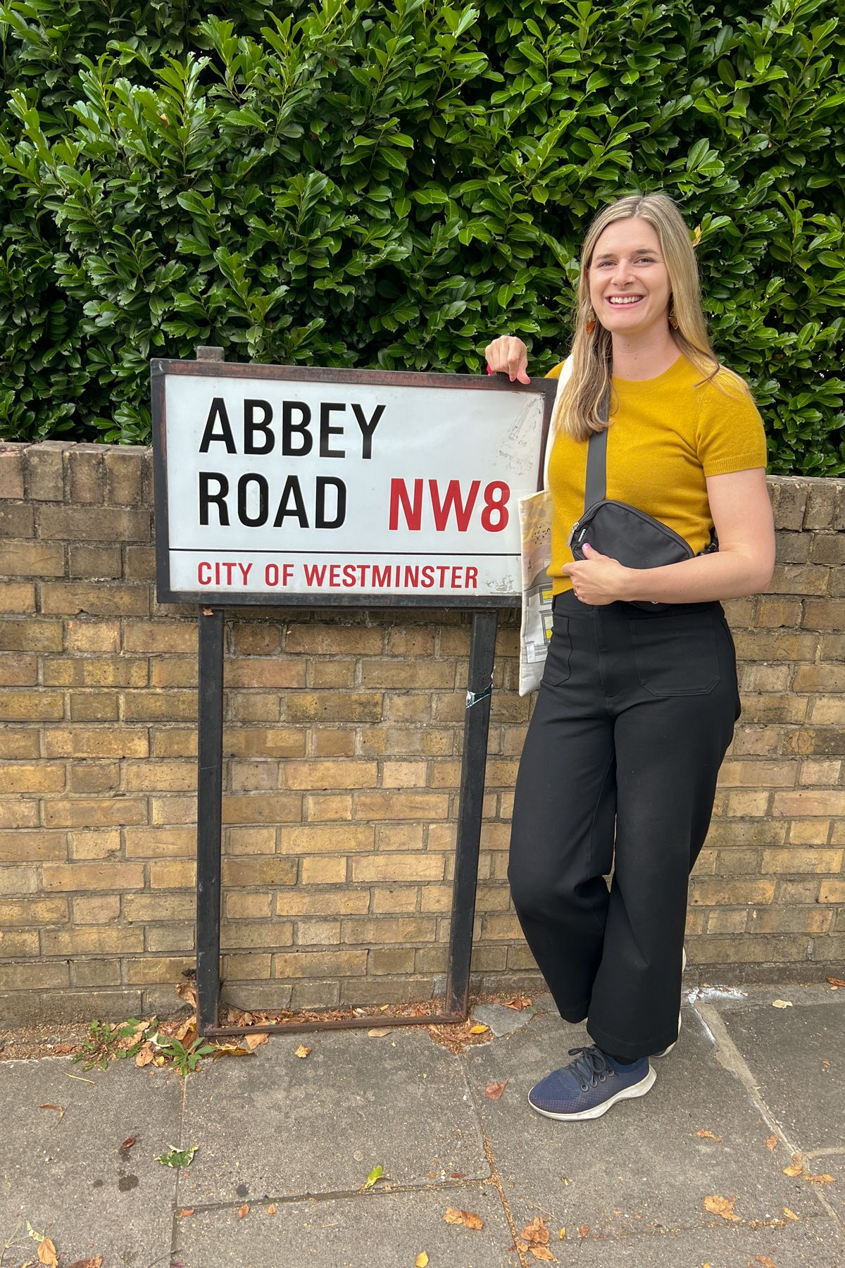 A young woman wearing black pants and a yellow Mongolian Cashmere Tee poses next to a sign reading "Abbey Road" in front of a brick wall.