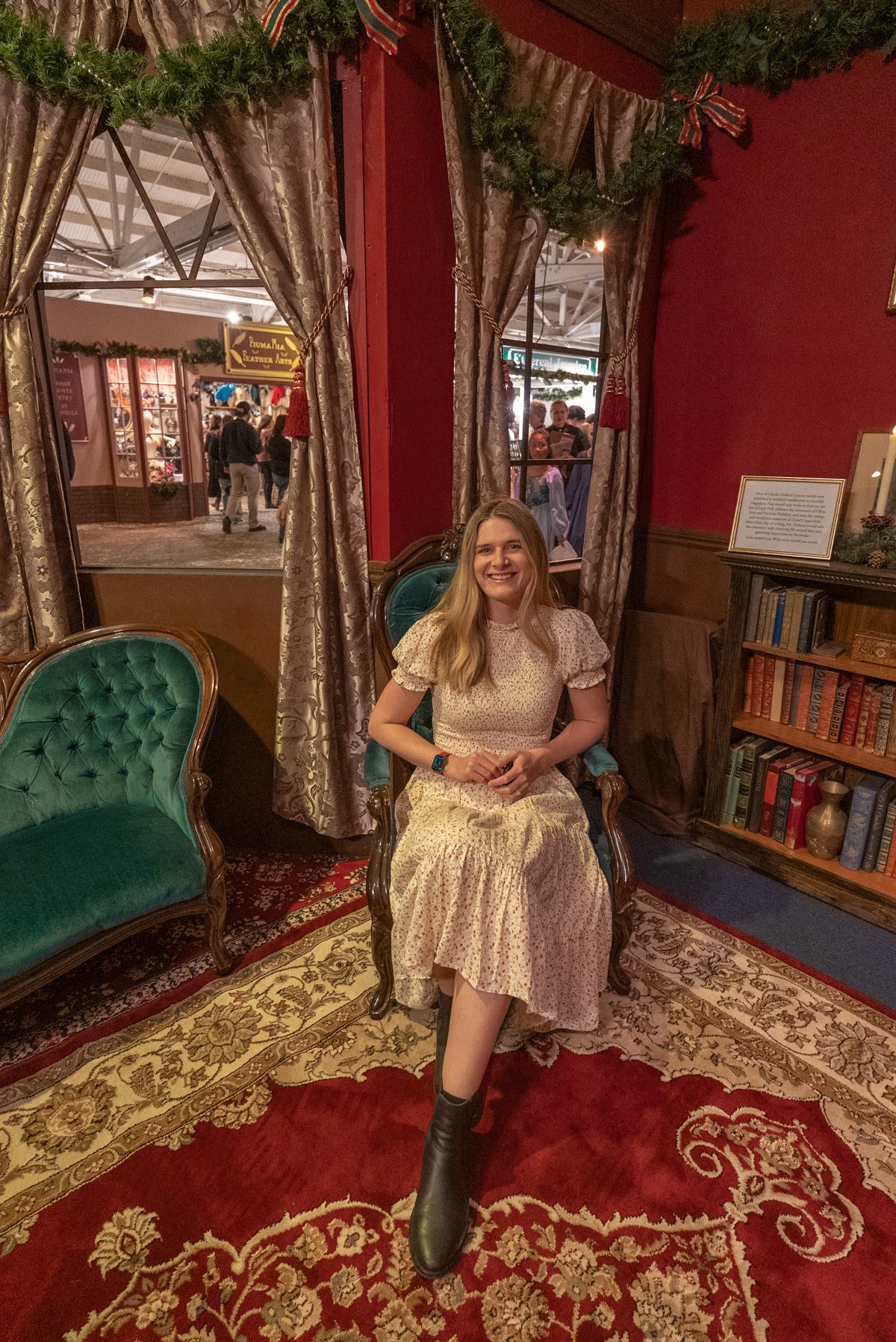 A woman in a white dress sits in an armchair in a red room decorated like a Victorian interior next to a green velvet couch and a bookshelf.