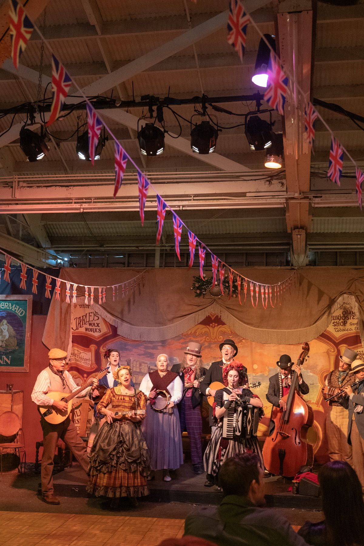 A crowd of costumed Dickensian performers sing together on a warmly-lit stage.