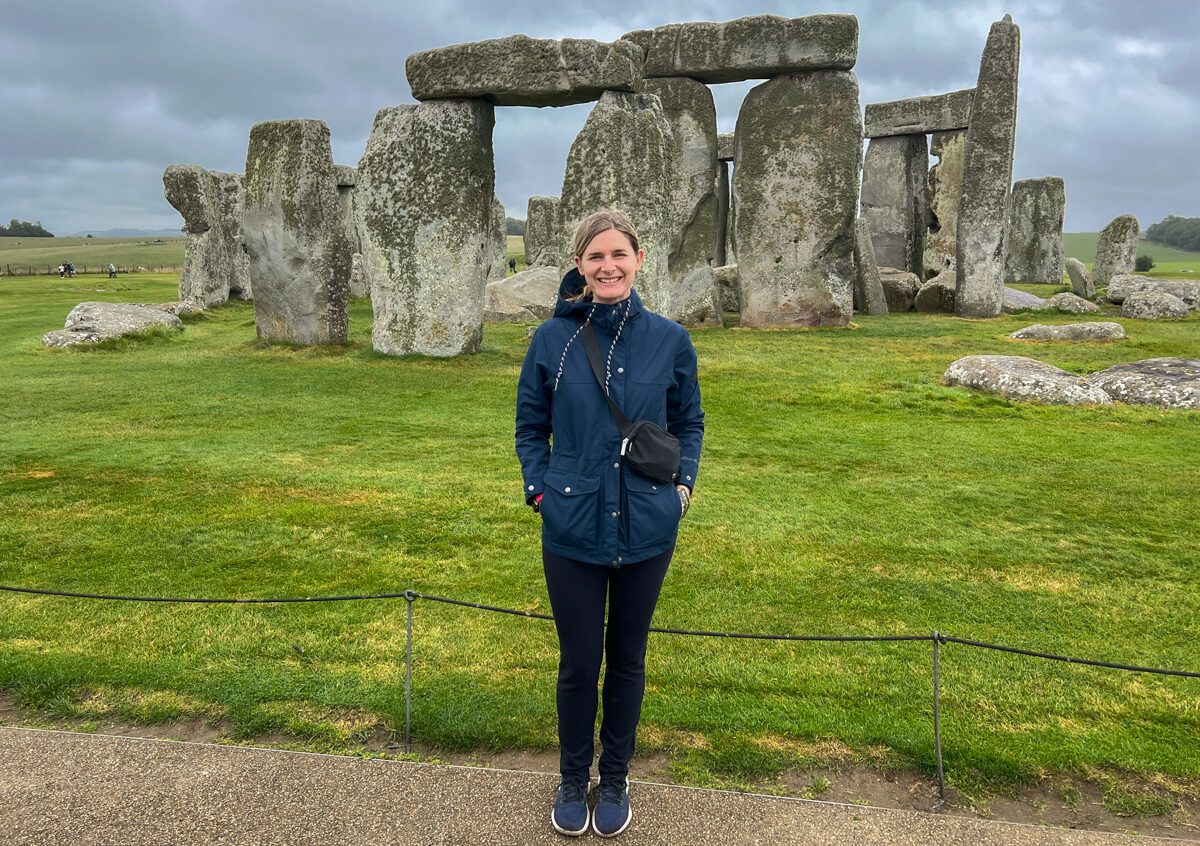 A young woman wearing black pants and a dark blue jacket, stands in front of Stonehenge in a rainy, hilly, green countryside landscape beneath an overcast sky.