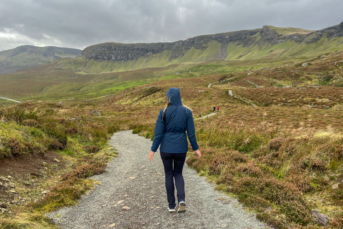 A young woman wearing black pants and a dark blue jacket walks down a road with her back to the camera in a rainy, hilly, green countryside landscape beneath an overcast sky.