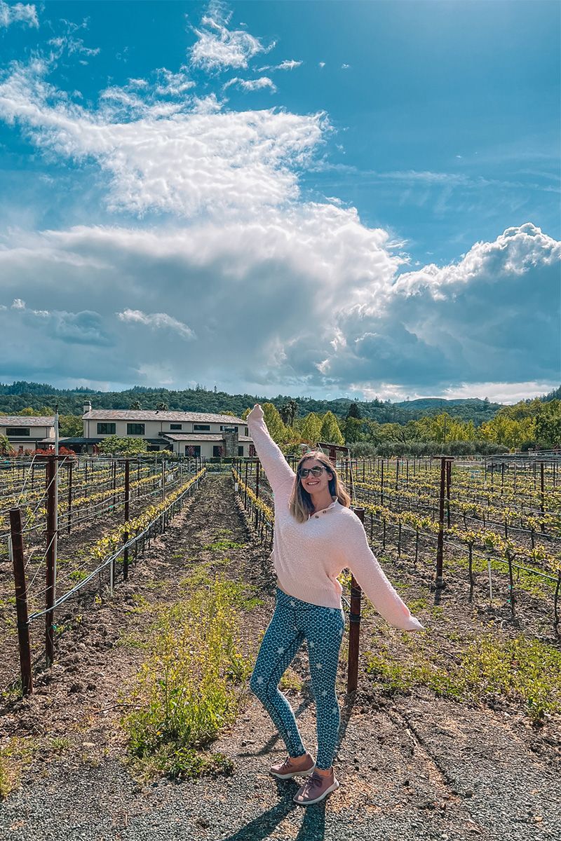 A woman wearing navy blue patterned leggings and a white sweater poses with one arm up in front of a vineyard on a sunny day.