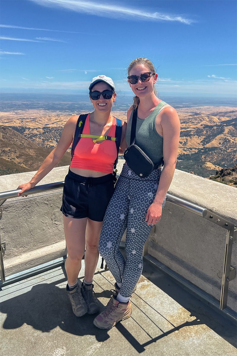 A woman wearing navy blue patterned leggings and a green tank top poses smiling next to a woman wearing black bike shorts and a pink sports bra on a scenic overlook with a valley and a blue sky behind them.