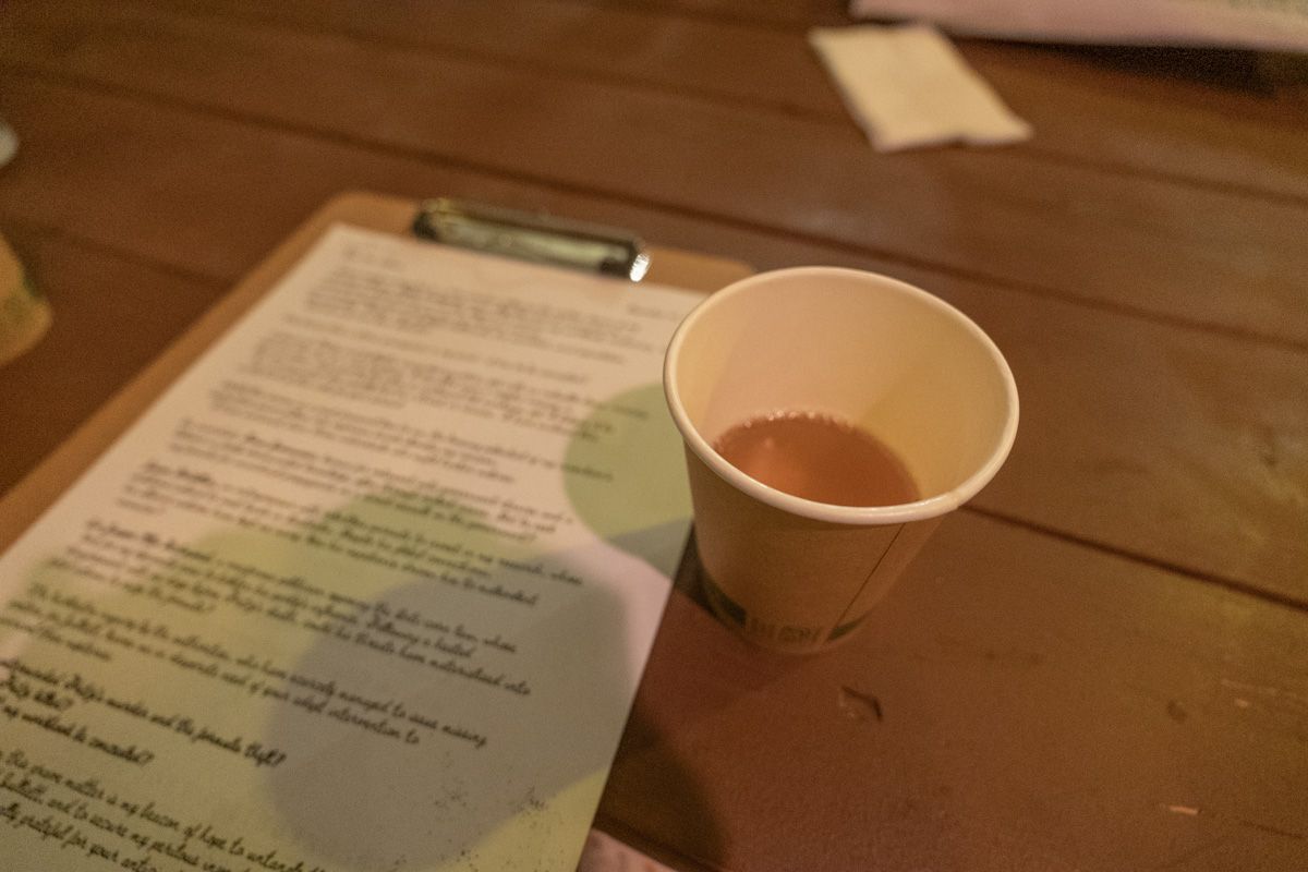 A paper cup containing amber liquid sits on a wooden table, with a clipboard covered in text in soft-focus beside it.