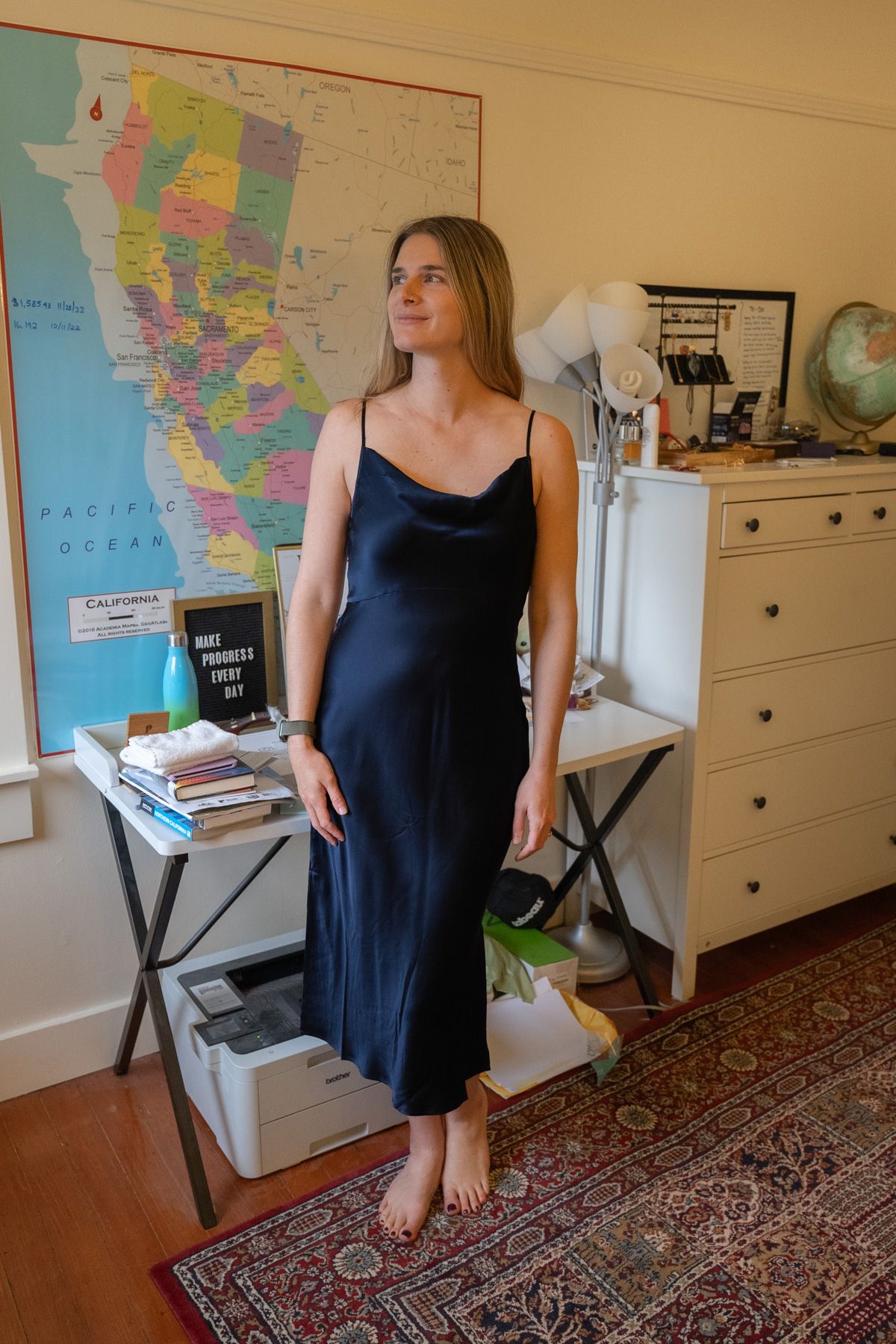 A young woman wearing a dark blue Silk Slip Dress stands looking over her right shoulder in an interior setting with a map of California on the wall behind her.