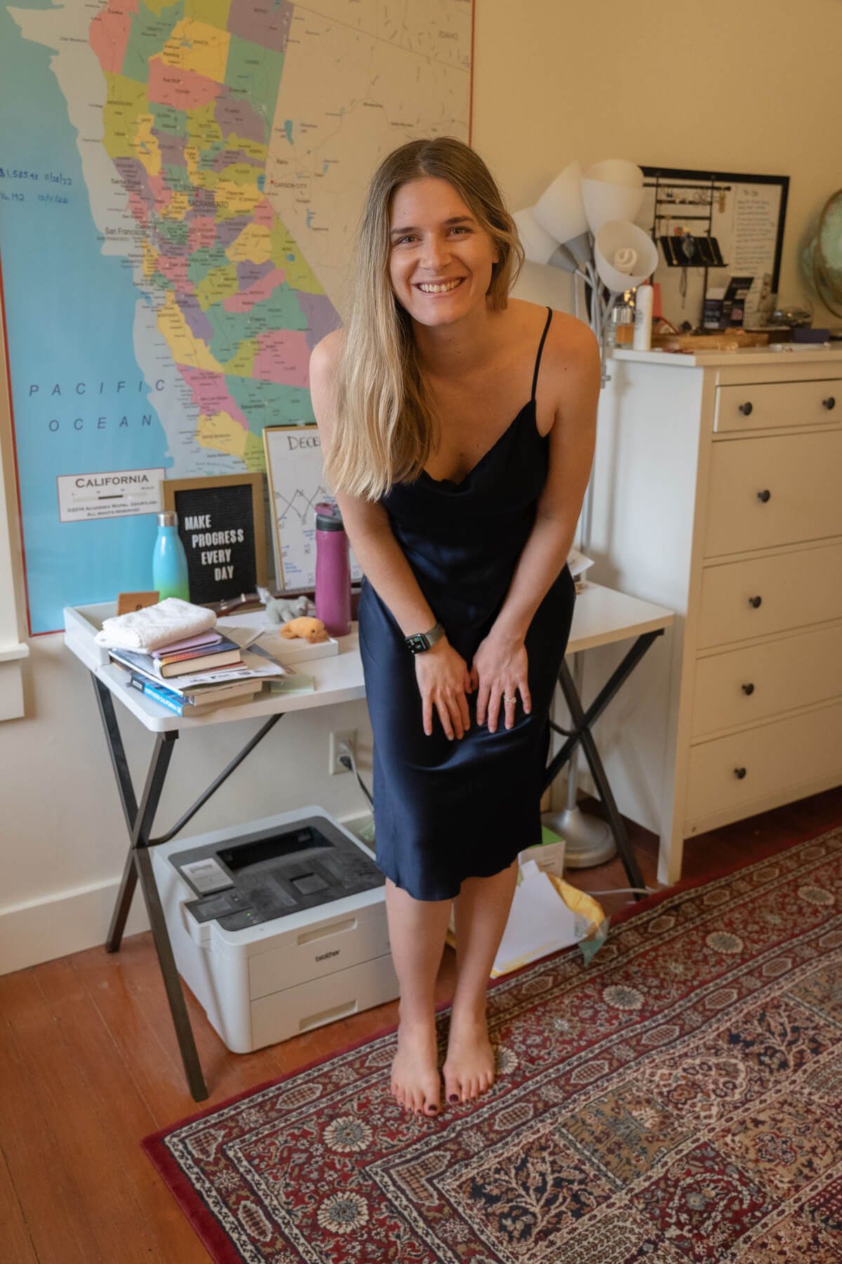 A young woman wearing a dark blue Silk Slip Dress smiles and leans forward with her hands on her knees in an interior setting with a map of California on the wall behind her.