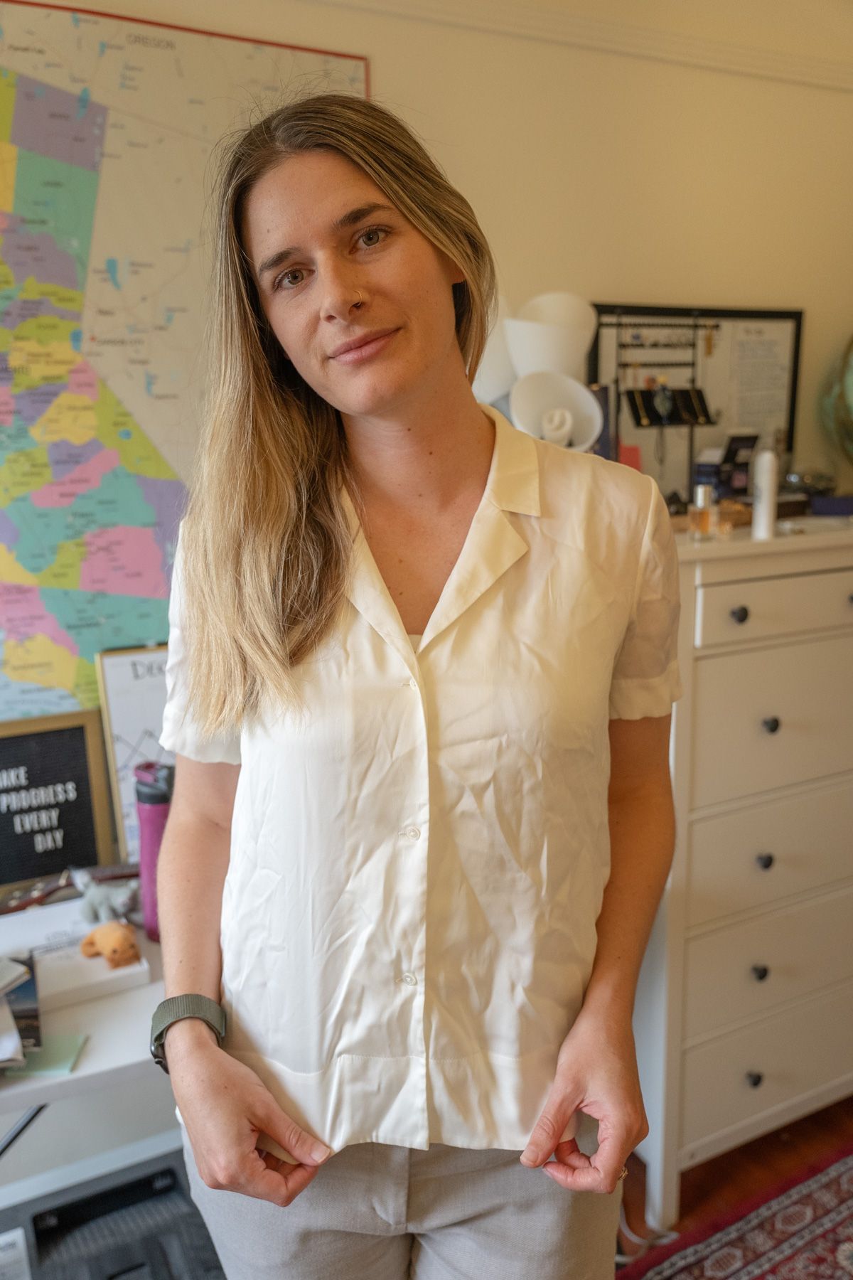 A young woman wearing a white Stretch Silk Short Sleeve Notch Collar Blouse stands, not smiling, in an interior setting with a map of California on the wall behind her.