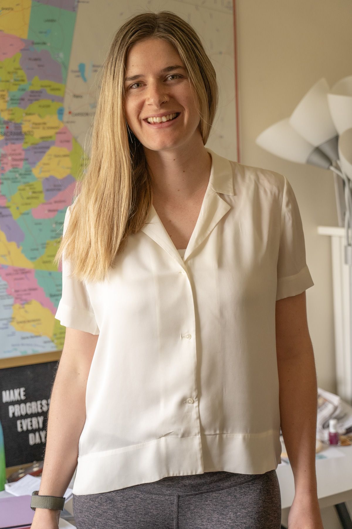 A young woman wearing a white Stretch Silk Short Sleeve Notch Collar Blouse stands facing the camera, smiling, in an interior setting with a map of California on the wall behind her.