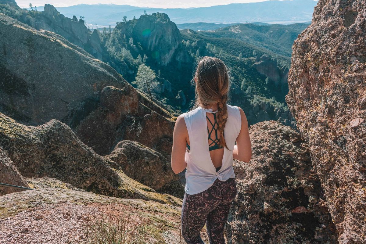 A woman wearing a white, open-back top and patterned leggings stands with her back to the camera looking out a mountainous landscape.