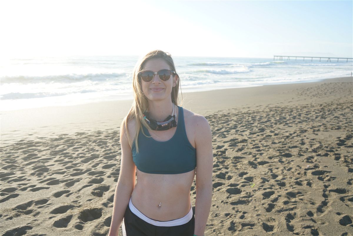 A woman wearing sunglasses and a teal sports bra stands on a beach and smiles at the camera.