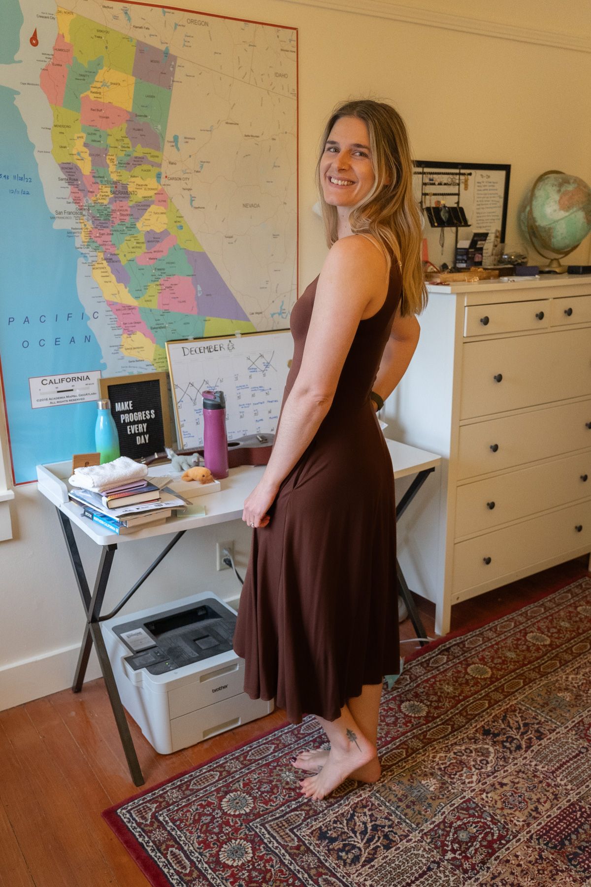 A young woman wearing a brown Tencel Jersey Fit & Flare Dress stands to the side, smiling over her shoulder in an interior setting with a map of California on the wall behind her.