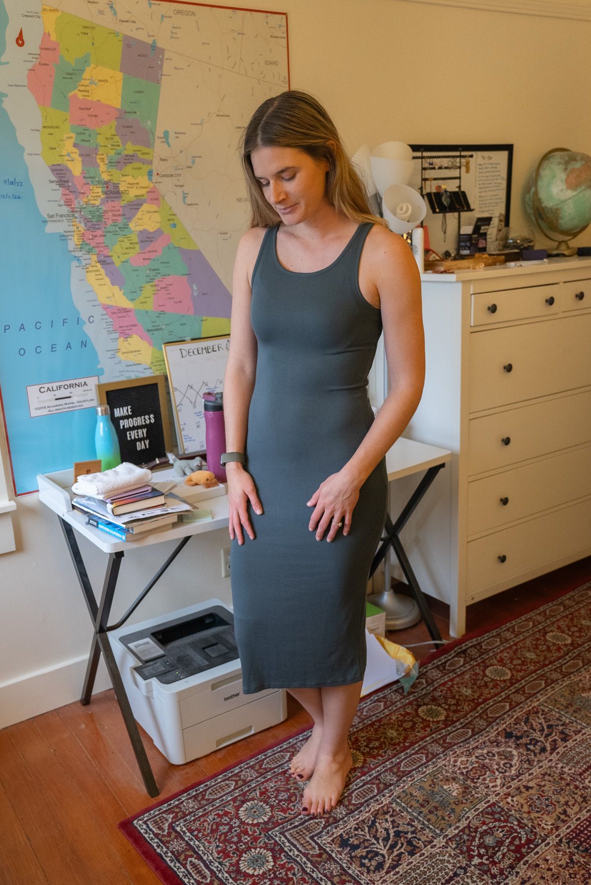 A young woman wearing a grey Tencel Rib Knit Sleeveless Dress stands looking down in an interior setting with a map of California on the wall behind her.
