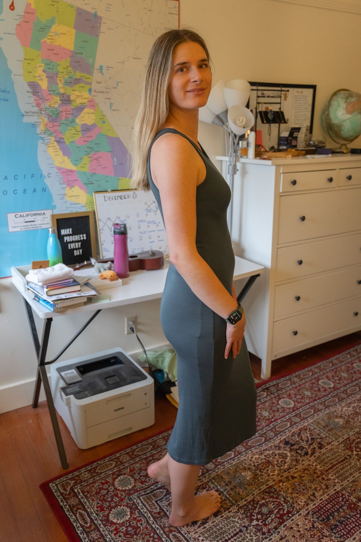 A young woman wearing a grey Tencel Rib Knit Sleeveless Dress stands to the side, smiling over her shoulder in an interior setting with a map of California on the wall behind her.