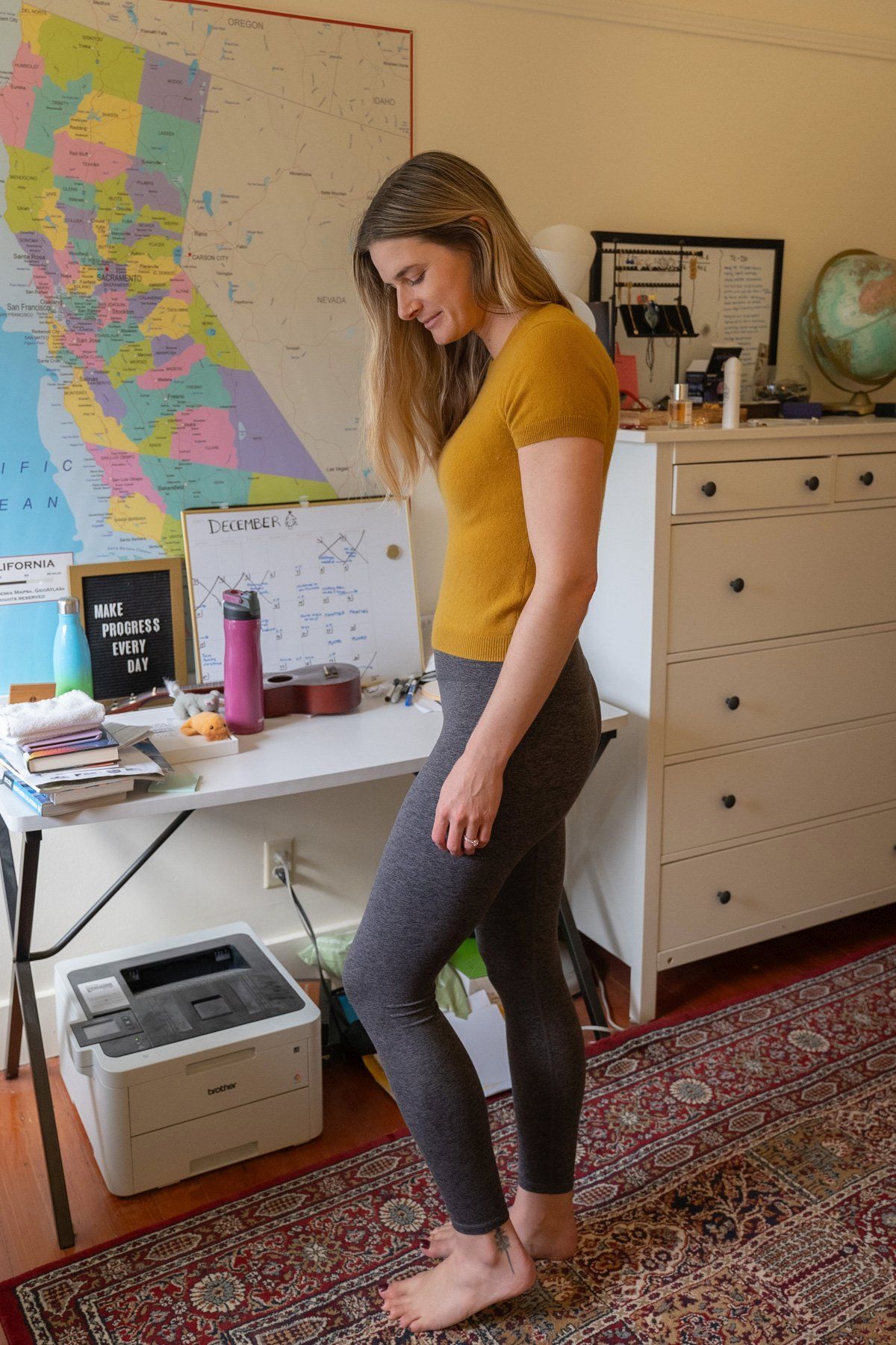 A young woman wearing grey Ultra-Soft High-Rise Leggings and a yellow shirt stands to the side, looking down in an interior setting with a map of California on the wall behind her.
