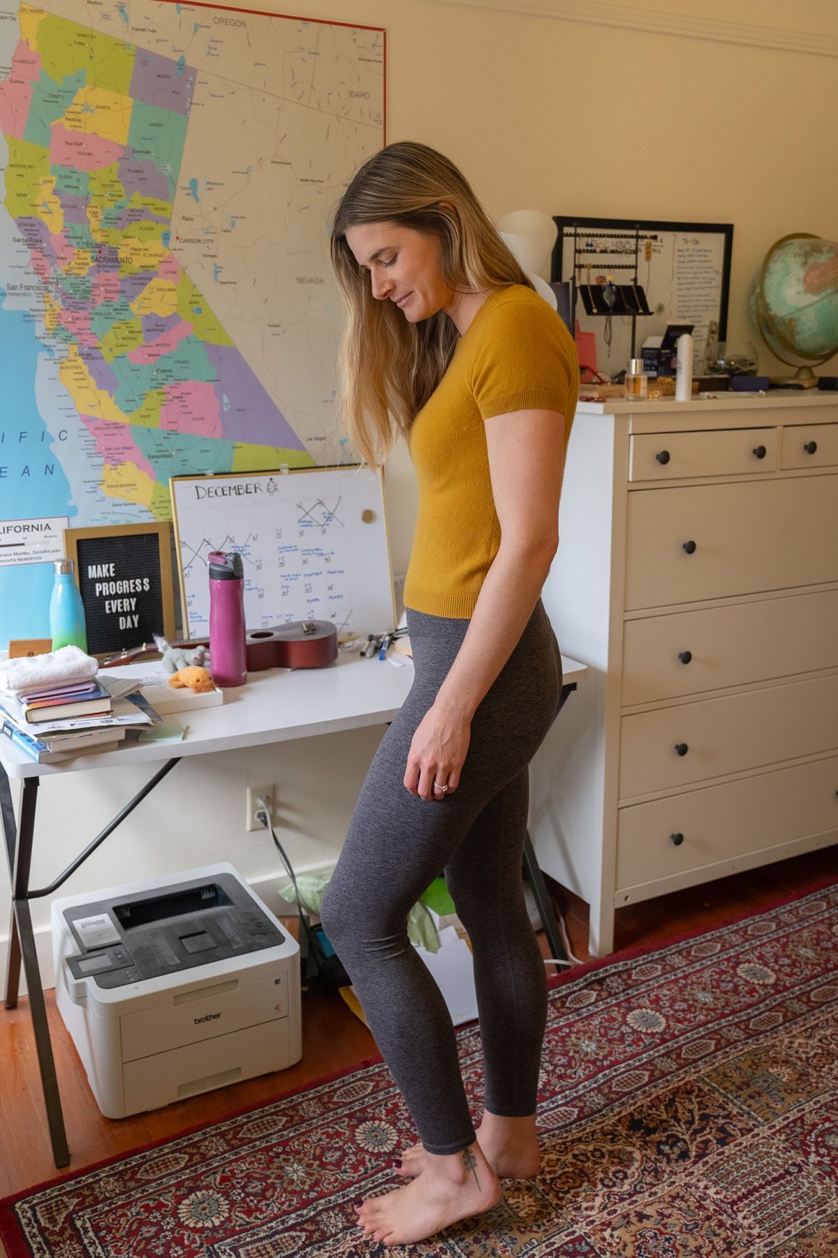 A young woman wearing grey Ultra-Soft High-Rise Leggings and a yellow shirt stands to the side, looking down in an interior setting with a map of California on the wall behind her.