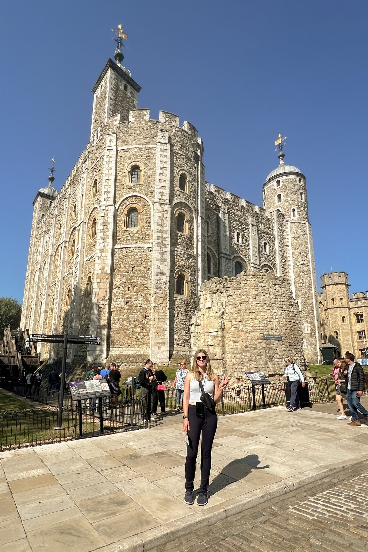 A young woman in black pants and a white tank top stands in front of the Tower of London on a sunny day.