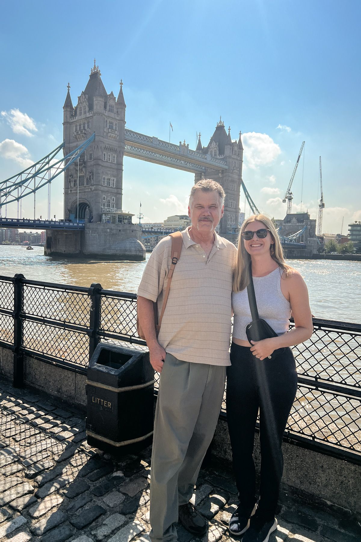 An older man in a white t-shirt stands next to a young woman in black pants and a white tank top in front of the London Bridge on a sunny day.