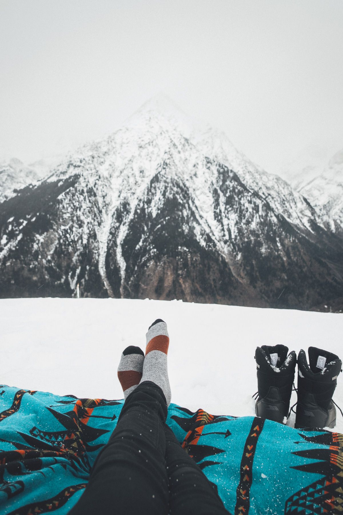 A POV-shot of a pair of feet wearing socks, lounging on a turquoise quilt on a snowy mountain-top, looking out at a wintry mountain landscape. 