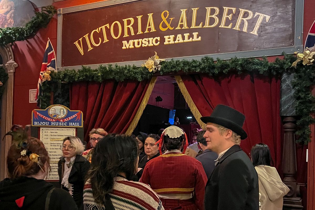 People in Dickensian costumes stand lined up in front of a doorway decorated with red velvet curtains and a Christmas garland, beneath a hand-painted sign that says, "Victoria & Albert Music Hall."
