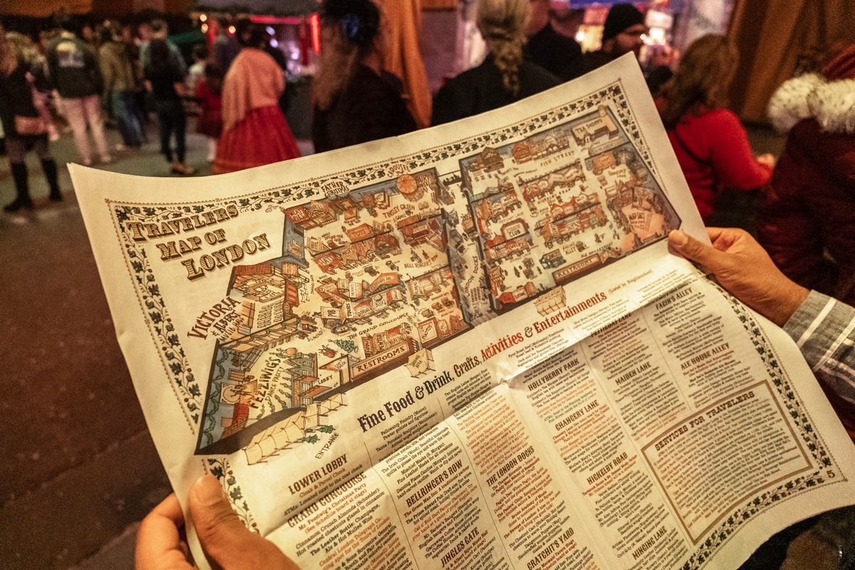 A pair of hands holding a map of the Dickens Fair, which is titled "Travelers Map of London," with crowds of costumed fairgoers in soft-focus in the background.