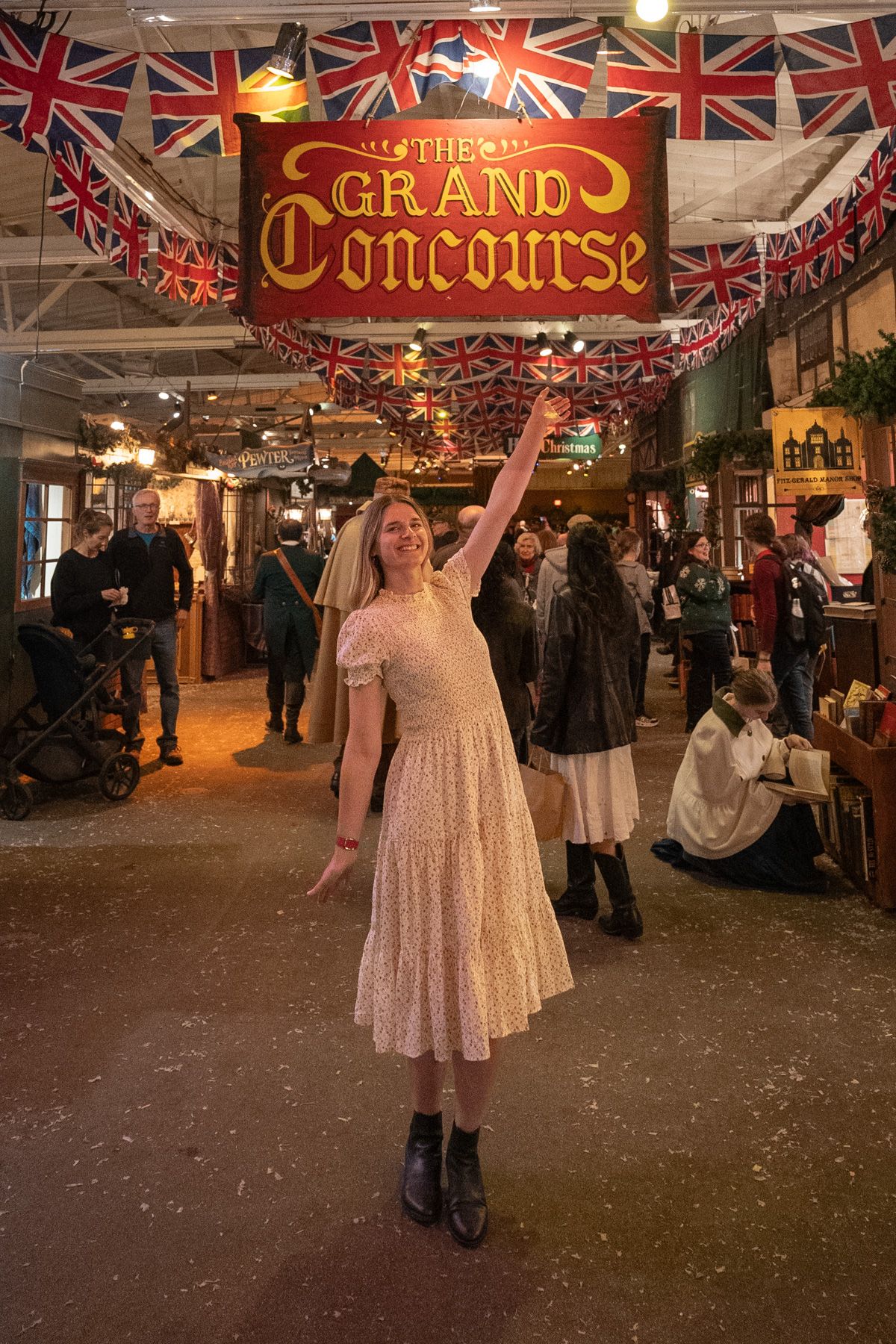 A woman in a white dress stands with her arm up gesturing towards a red and gold Grand Concourse sign at the Dickens Fair in San Francisco.