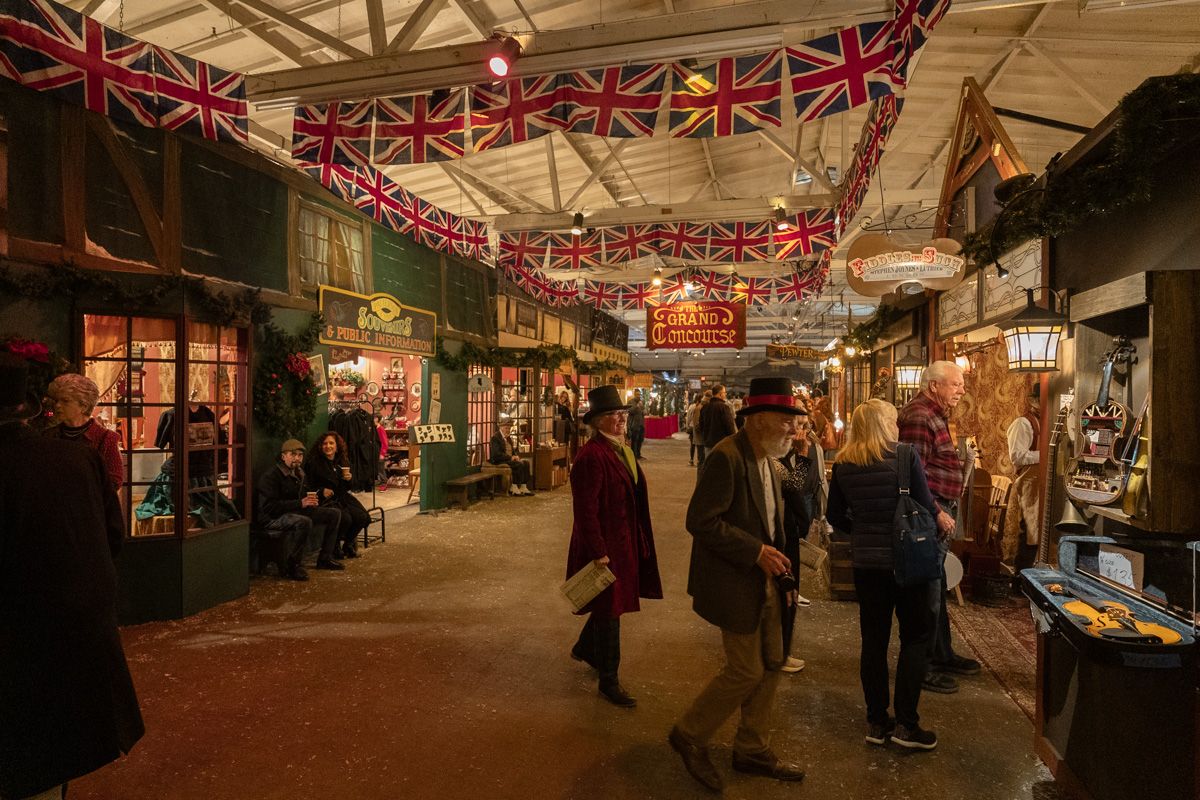 Costumed fairgoers mill about an indoor fair resembling a street in Victorian London, with garlands of British flags strung from the ceiling. 