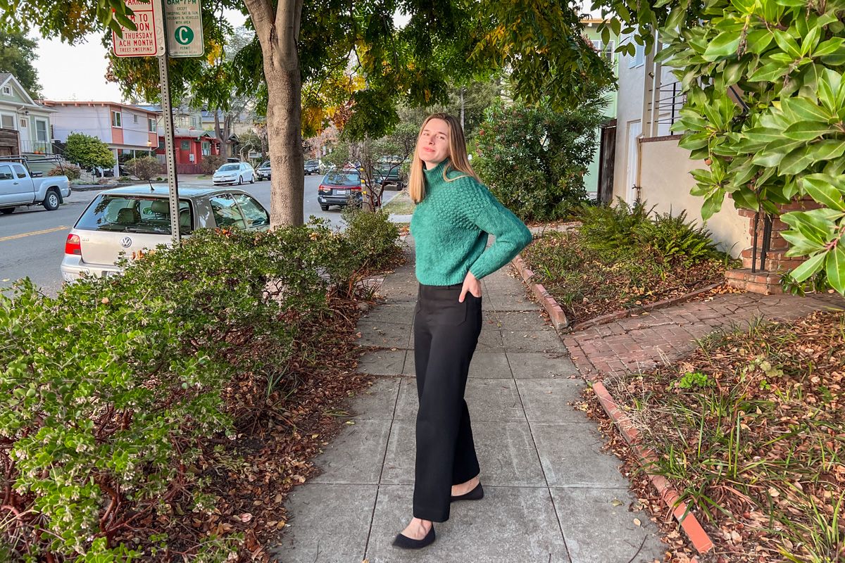A young, light-haired woman wearing a green sweater and black pants smiles at the the camera on a residential sidewalk, with shrubbery and trees behind her. 