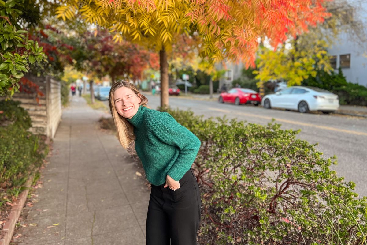 A young, light-haired woman wears a green sweater and black pants and laughs with her hands in her pockets on a residential sidewalk, with yellow autumn leaves surrounding her.