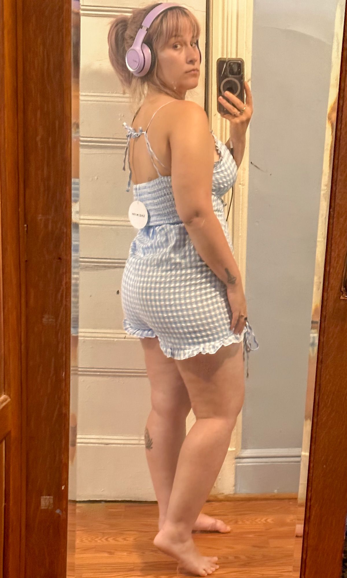 A purple-haired woman wearing a blue gingham romper takes a mirror selfie in a bedroom mirror.
