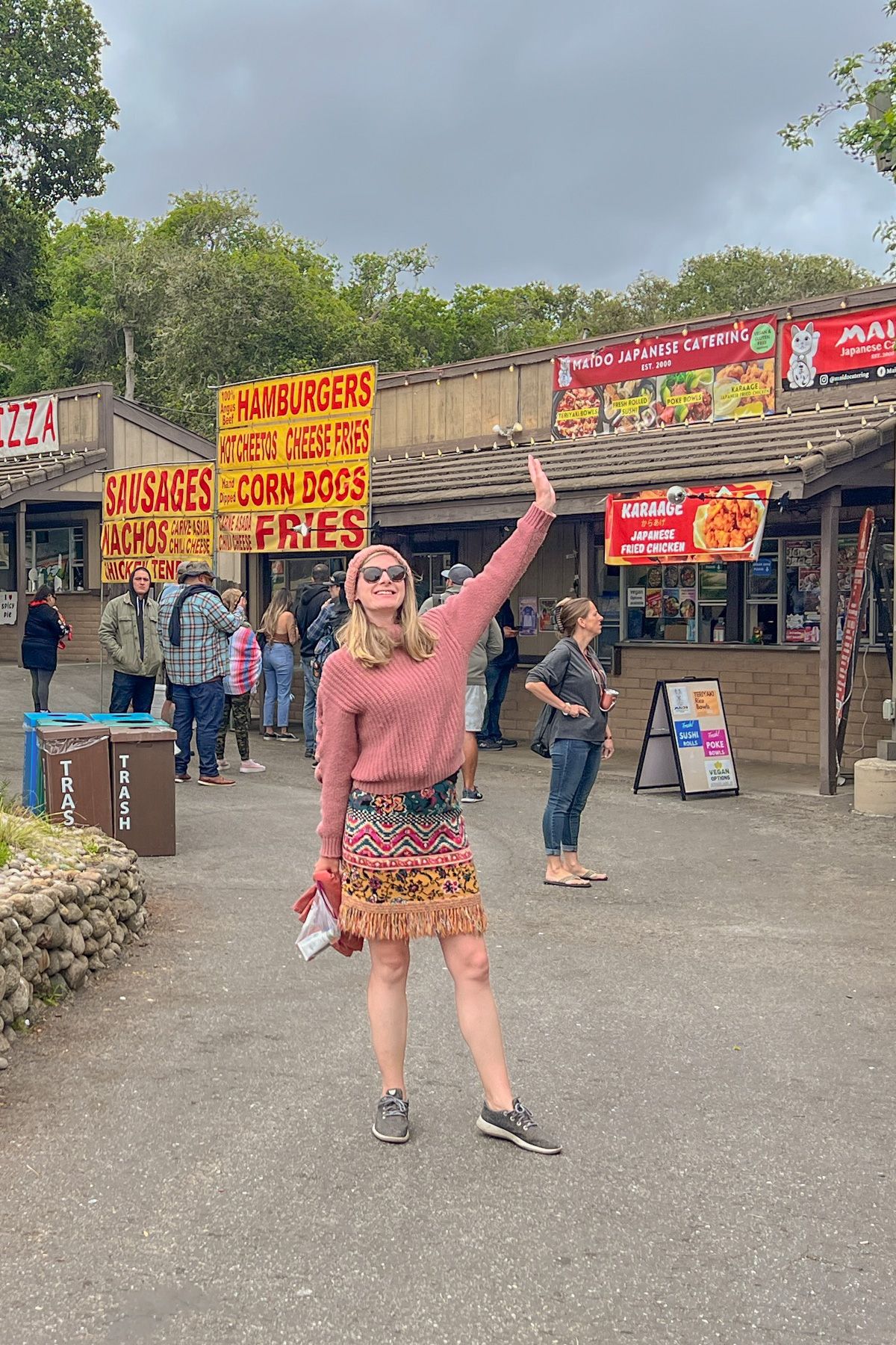 A young woman wearing a pink turtleneck sweater and a printed mini-skirt poses with one arm up in front of a row of fast food stands on a cloudy day.