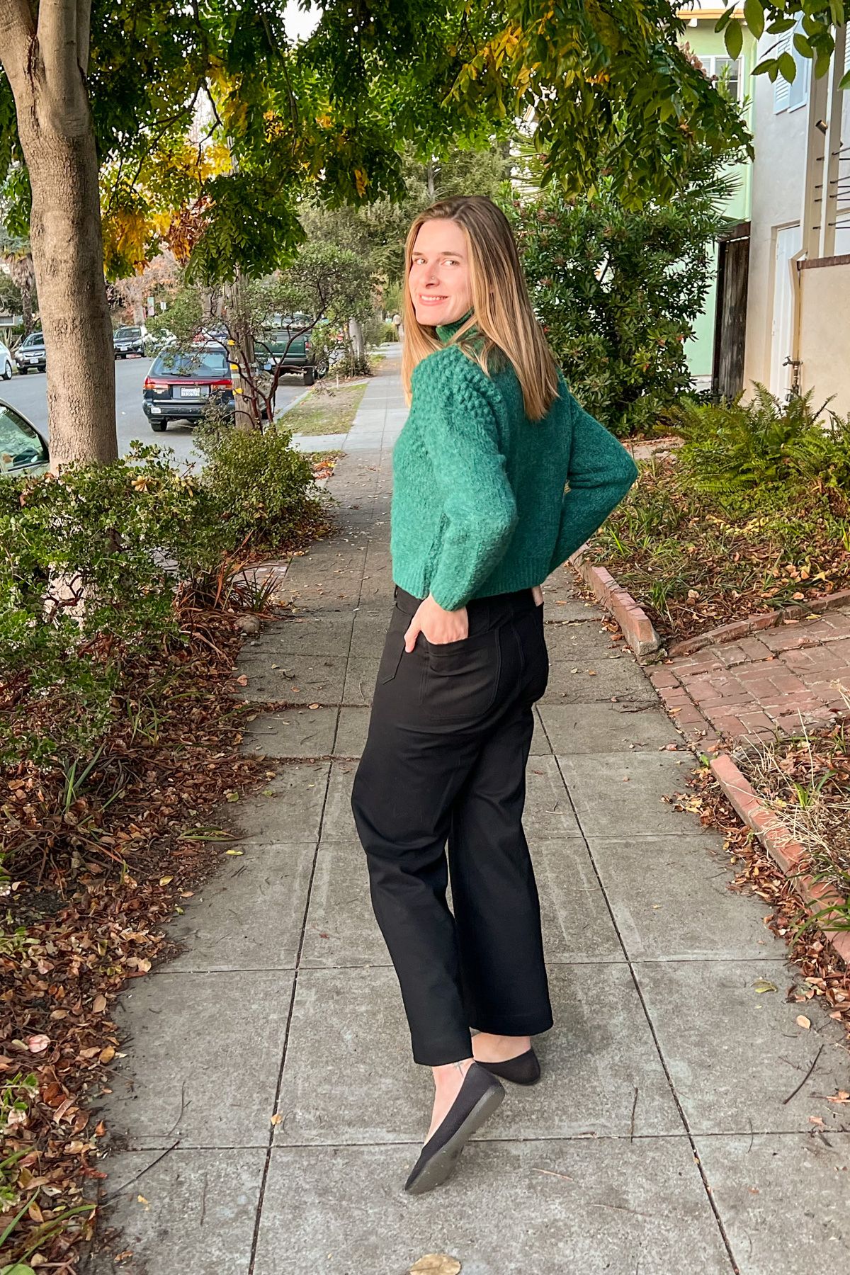 A young, light-haired woman wears a green sweater and black pants and poses with her hands in her back pockets, smiling over her shoulder on a residential sidewalk, with yellow autumn leaves surrounding her.