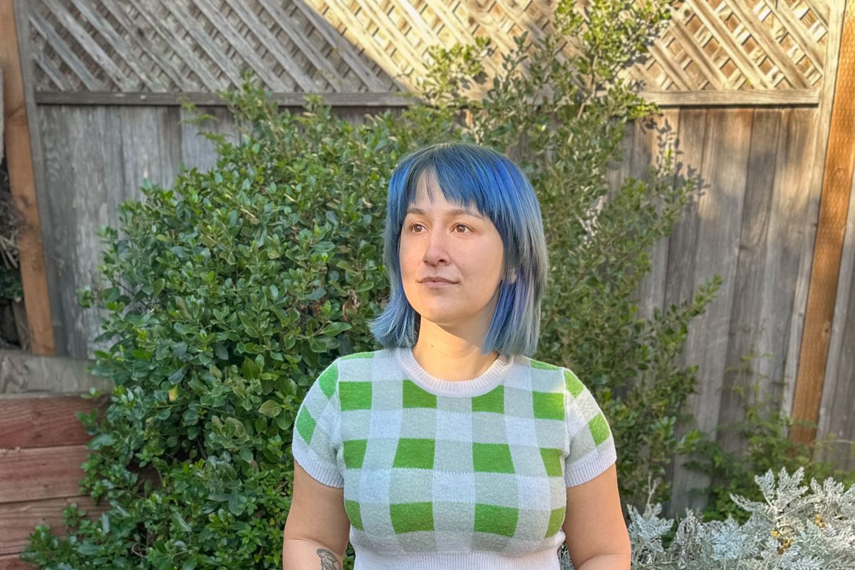 A blue-haired woman wearing a cropped, green and white checkered sweater looks into the distance in front of a backyard fence.