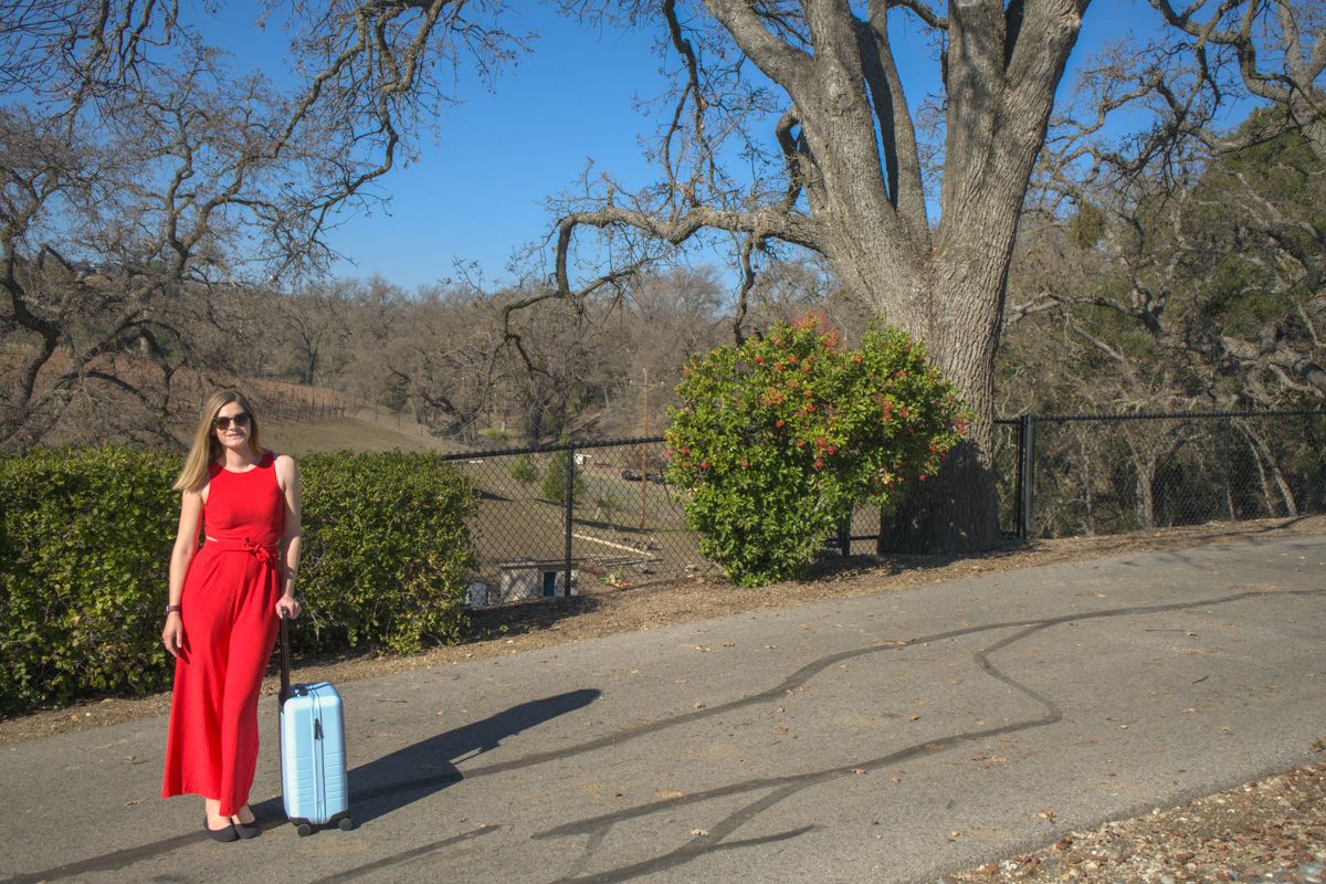 A young woman in a red romper stands with a light blue suitcase on a driveway on a sunny day, with trees and rolling hills behind her.