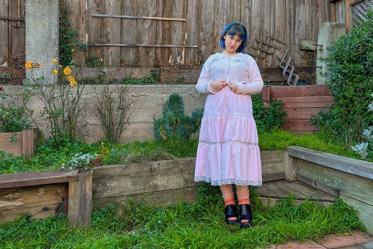 A blue-haired woman wearing a long, pink frilly skirt and matching cardigan, stands on a patch of grass in front of a backyard fence, with her hands clasped in front of her.