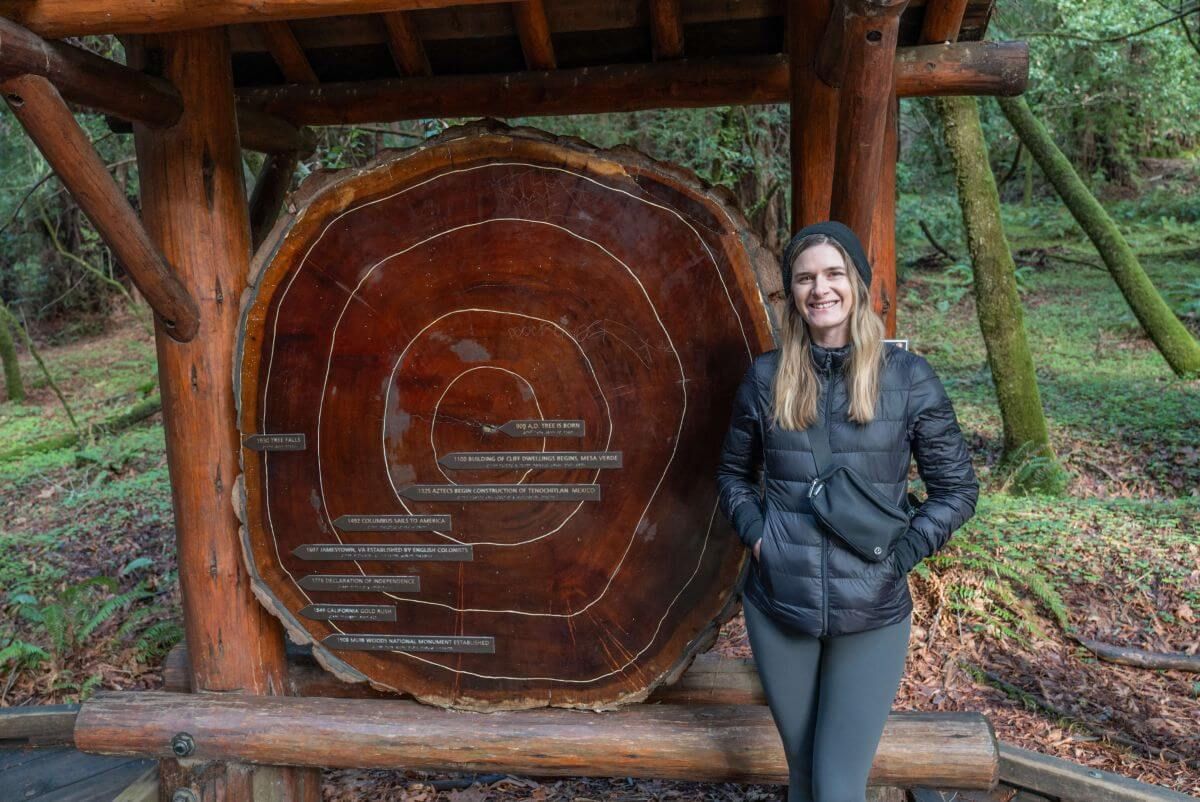 A woman wearing a black down jacket smiles at the camera, standing next a trailside display of a redwood tree cross section.