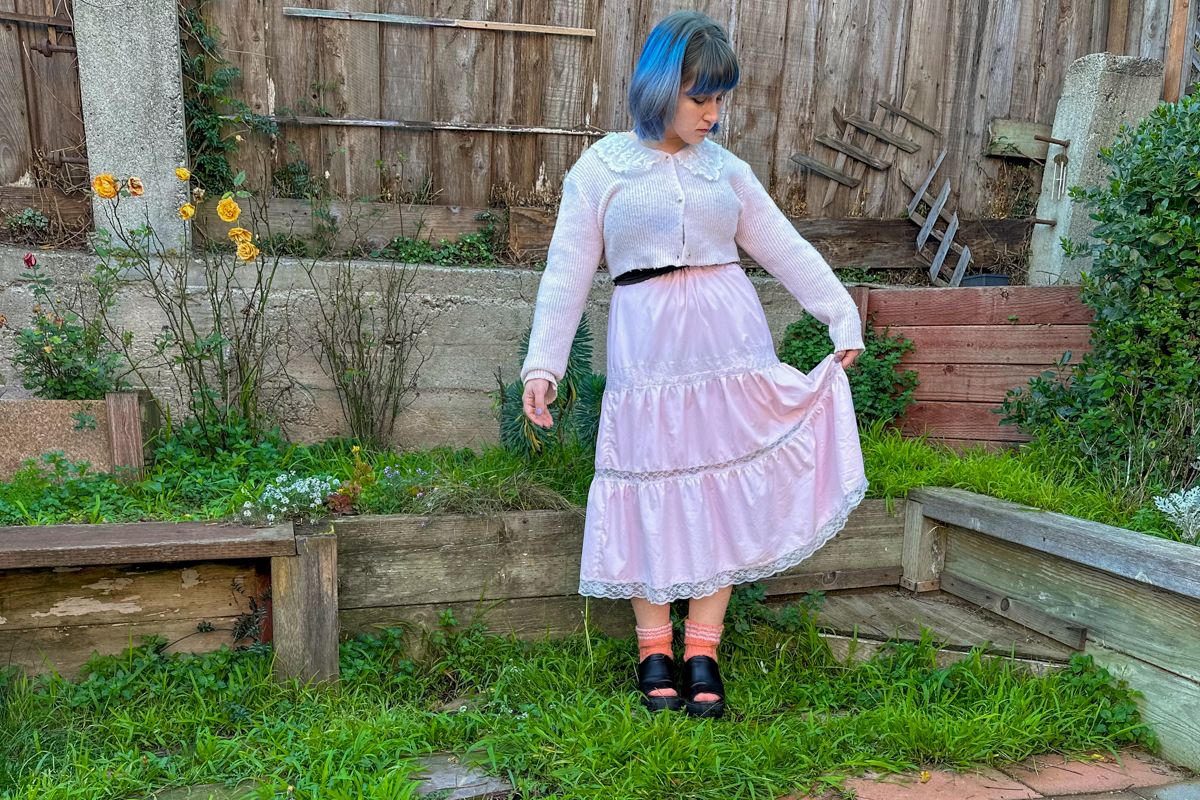 The author of this Romwe Review, a blue-haired woman wearing a long, pink frilly skirt and matching cardigan, stands on a patch of grass in front of a backyard fence.
