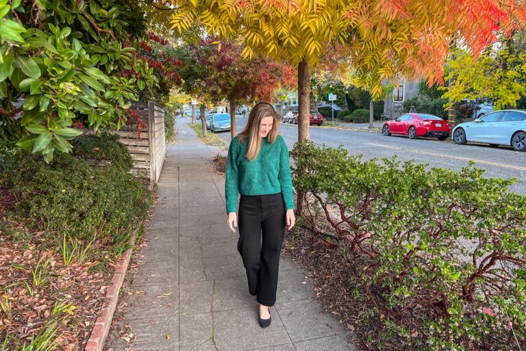 The author of this Nuuly Review, a young, light-haired woman, wears a green sweater and black pants while walking towards the camera on a residential sidewalk, with yellow autumn leaves surrounding her.