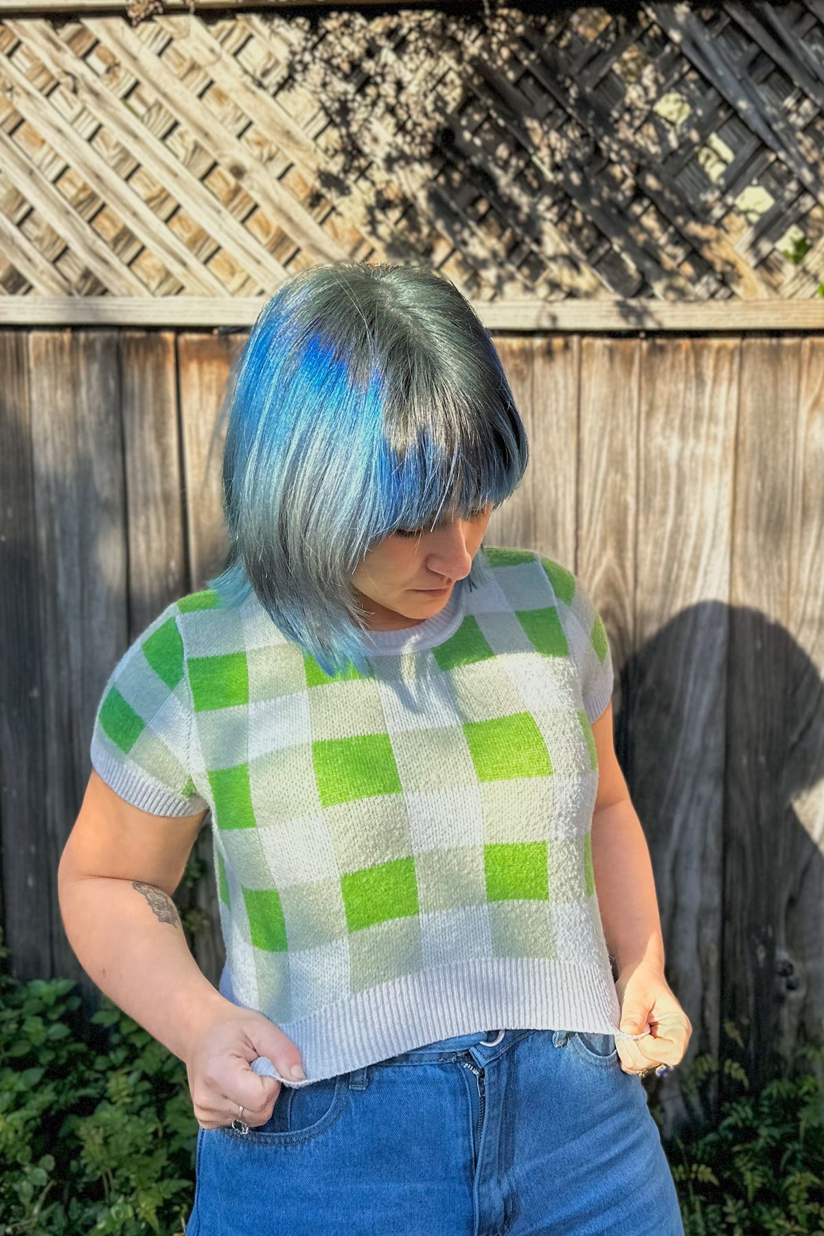 A blue-haired woman tugs down her cropped, green and white checkered sweater while standing in front of a backyard fence.