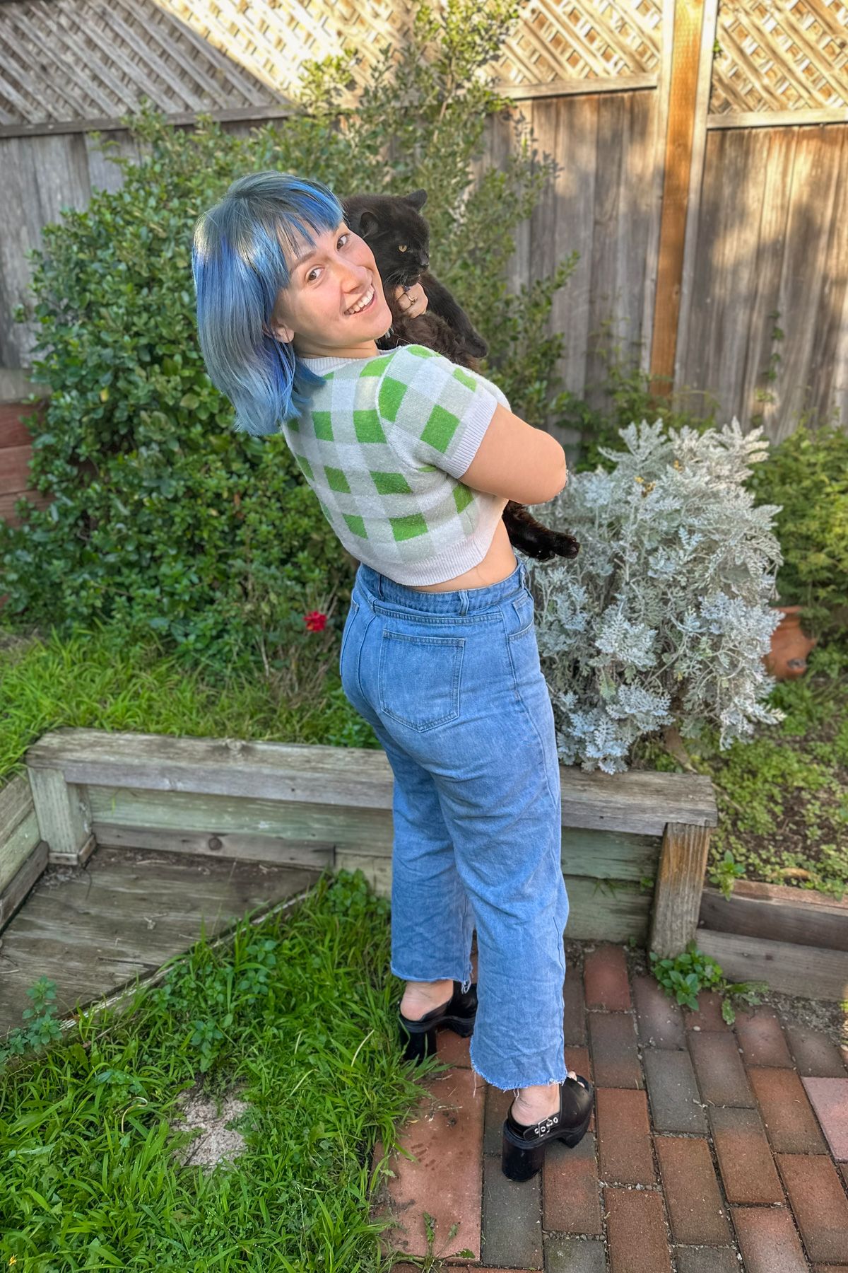 A blue-haired woman wearing a cropped, green and white checkered sweater and jeans smiles over her shoulder, holding a black cat, while standing on a brick patio in front of a backyard fence.