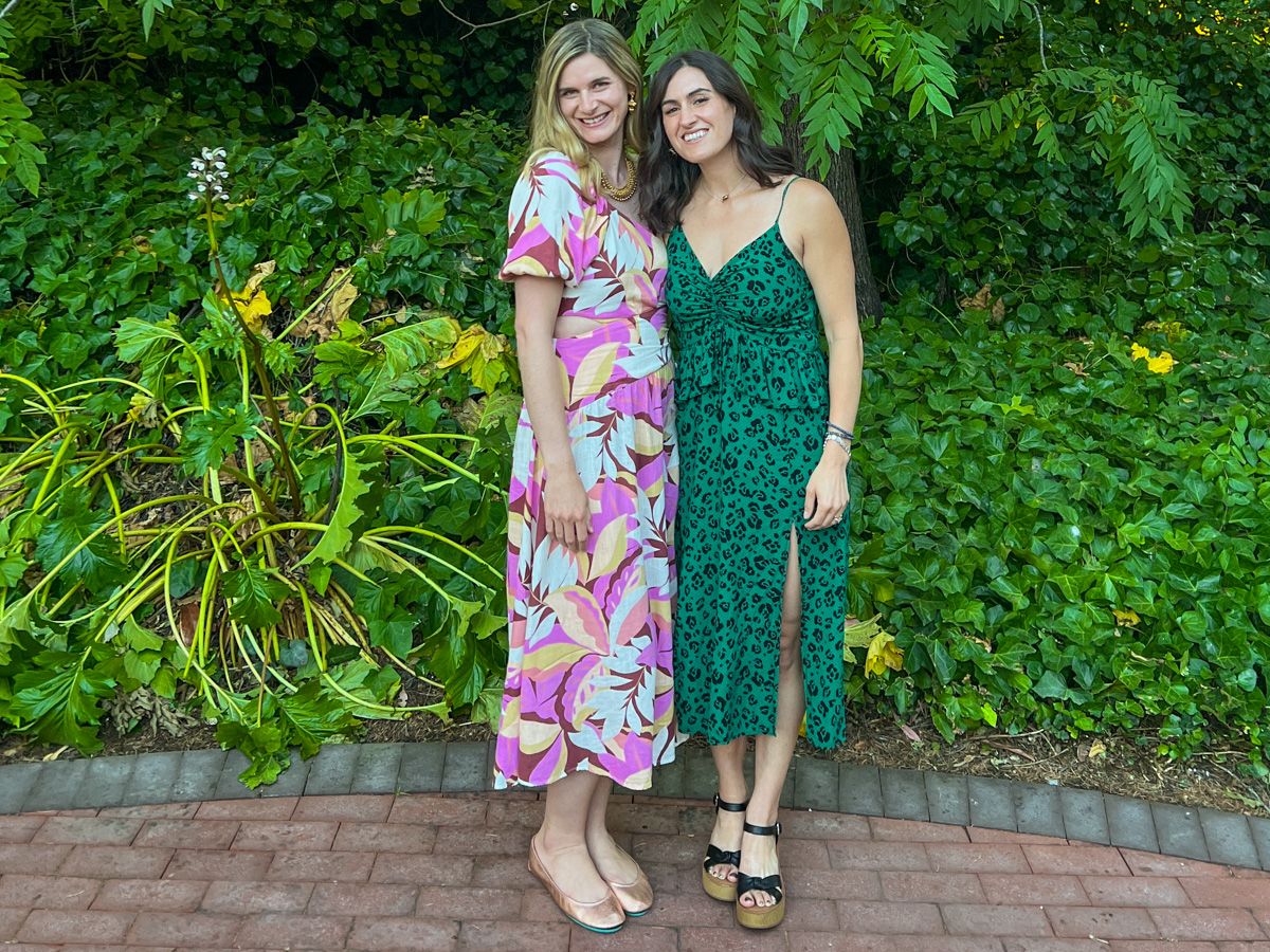 Two young women, one wearing a pink and white tropical-print dress, the other in a green cheetah-print one, pose side-by-side in front of a hill of greenery on a brick patio.