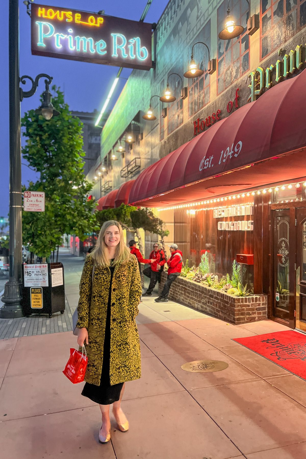 A young, light-haired woman wears a yellow leopard-print wool coat and holds a red purse and stands facing the camera in front of House of Prime Rib on a city sidewalk at dusk.