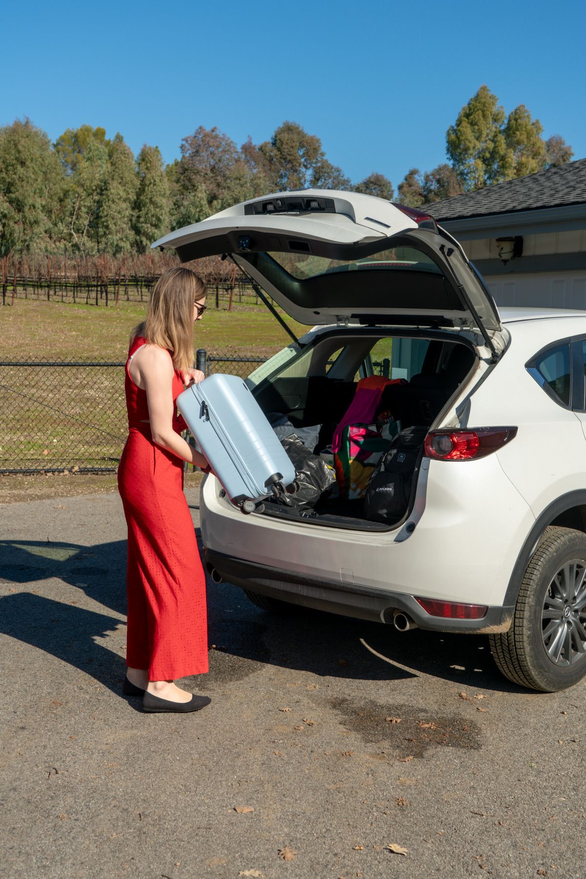 A woman in a red jumpsuit loads a blue suitcase into the hatchback of a white SUV parked in a driveway on a sunny day.