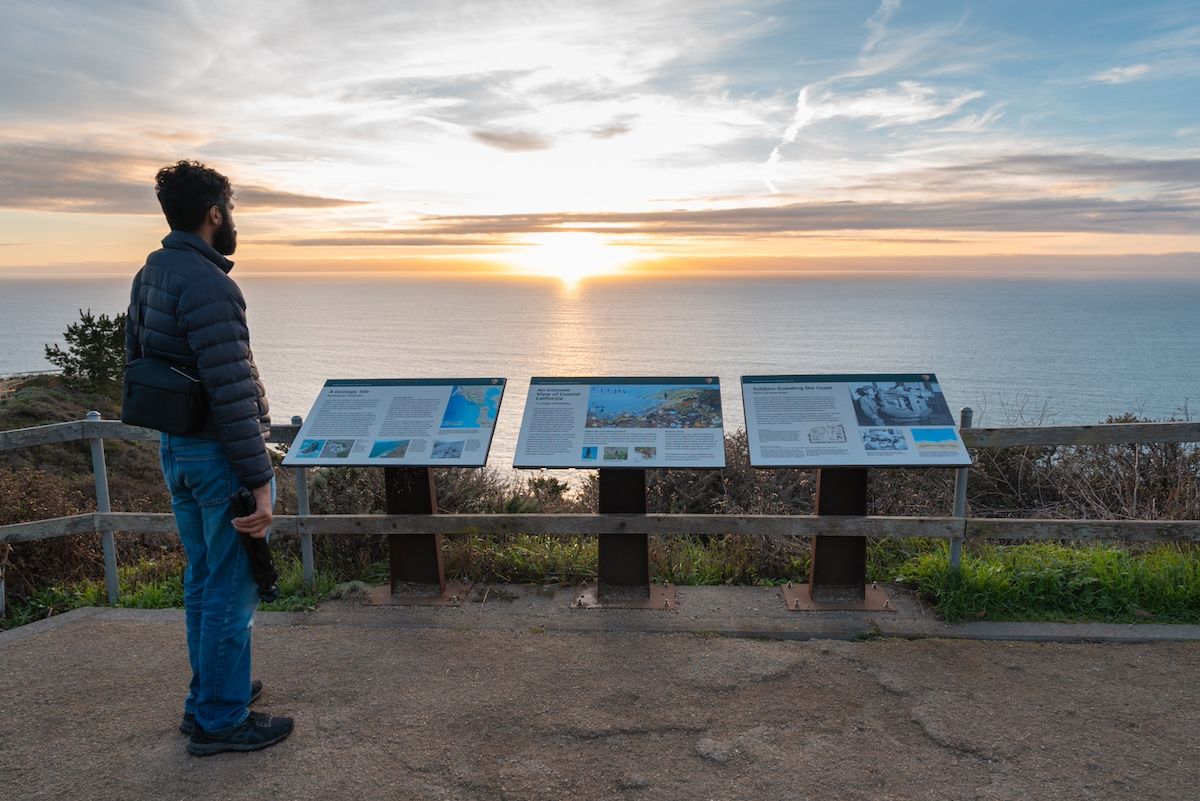 A man in jeans and windbreaker looking down at some informational plaques at the Muir Beach Overlook, bathed in the golden light of sunset.