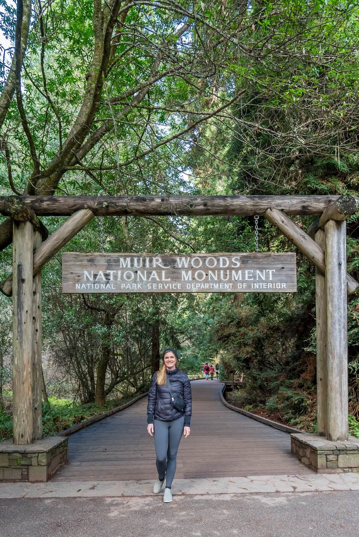 A woman stands under a wooden sign marking the entrance to Muir Woods National Monument, smiling at the camera.