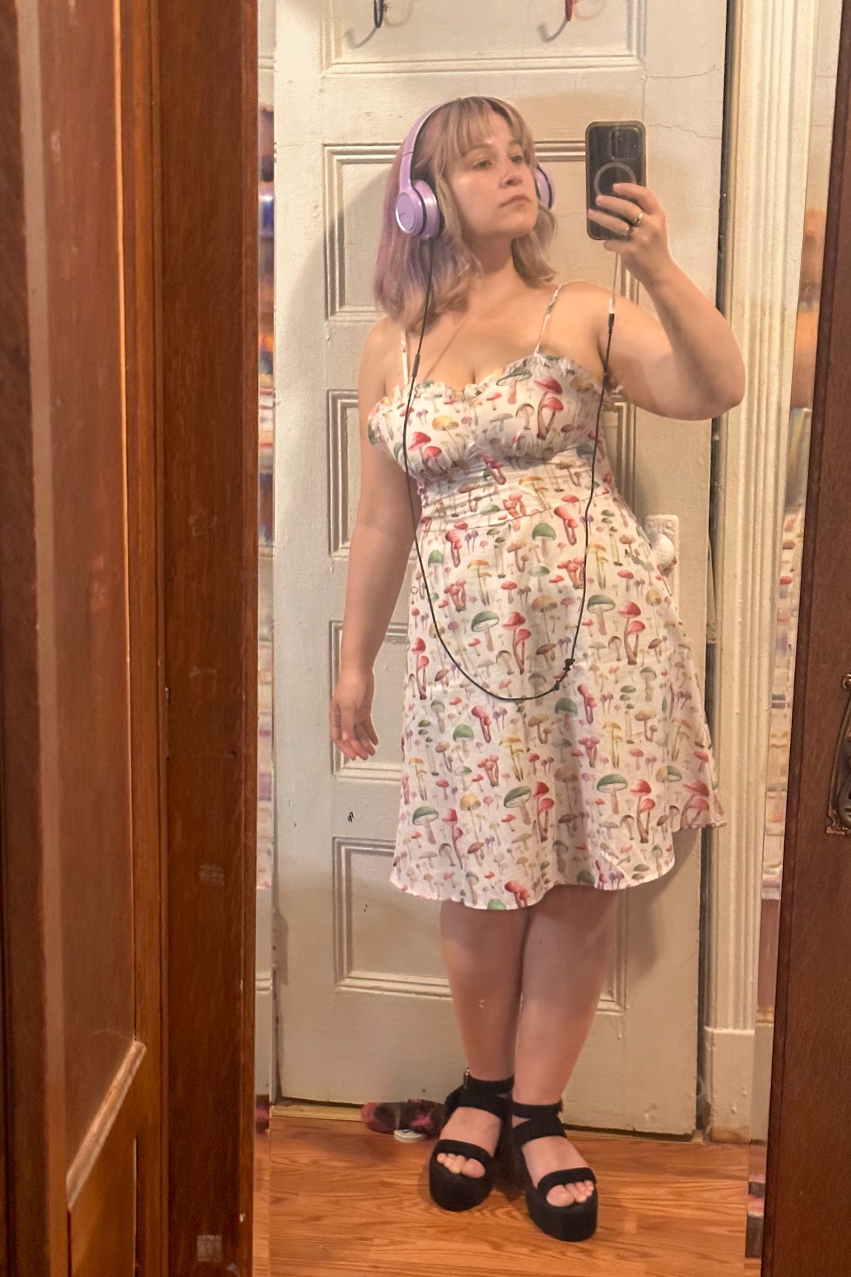A purple-haired woman wearing a mushroom-printed sundress takes a mirror selfie in a bedroom mirror.