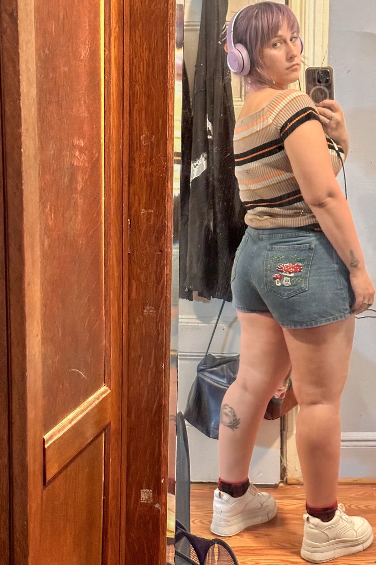 A purple-haired woman wearing jean shorts with a mushroom embroidered on the back pocket and a striped t-shirt takes a mirror selfie over her shoulder in a bedroom mirror.