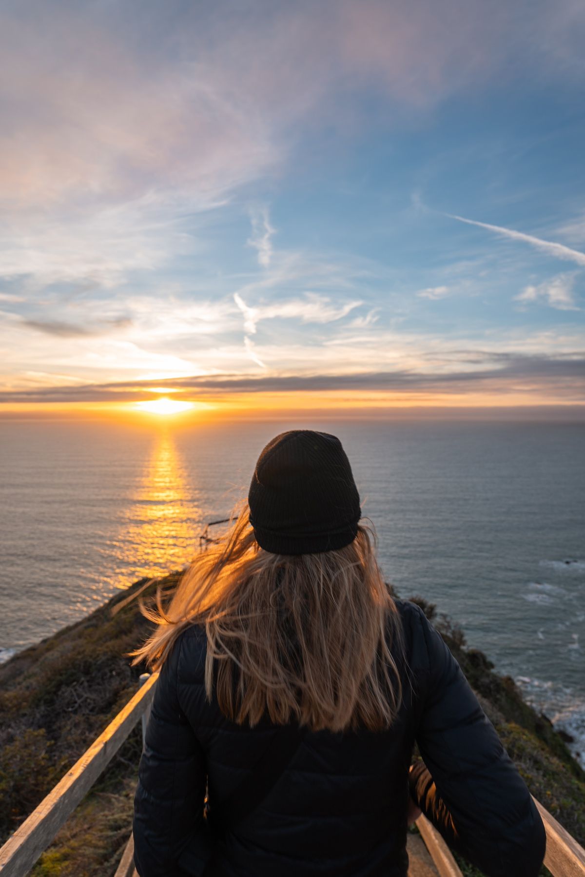 A view looking over the shoulder of a woman wearing a black beanie, looking out towards the Muir Beach Overlook, bathed in the golden light of sunset.